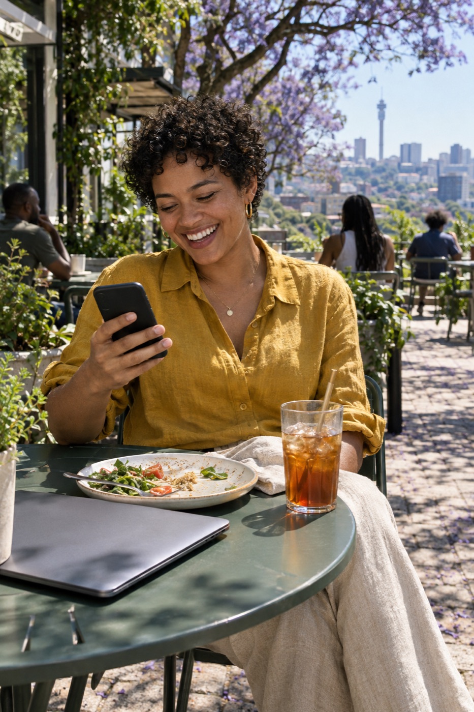 A woman laughing at her phone over lunch on a sunny café terrace, her laptop closed beside her